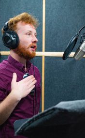 Expressive face of bearded man with red curly hair wear headphones near microphone who makes professional dubbing on a voice recording studio